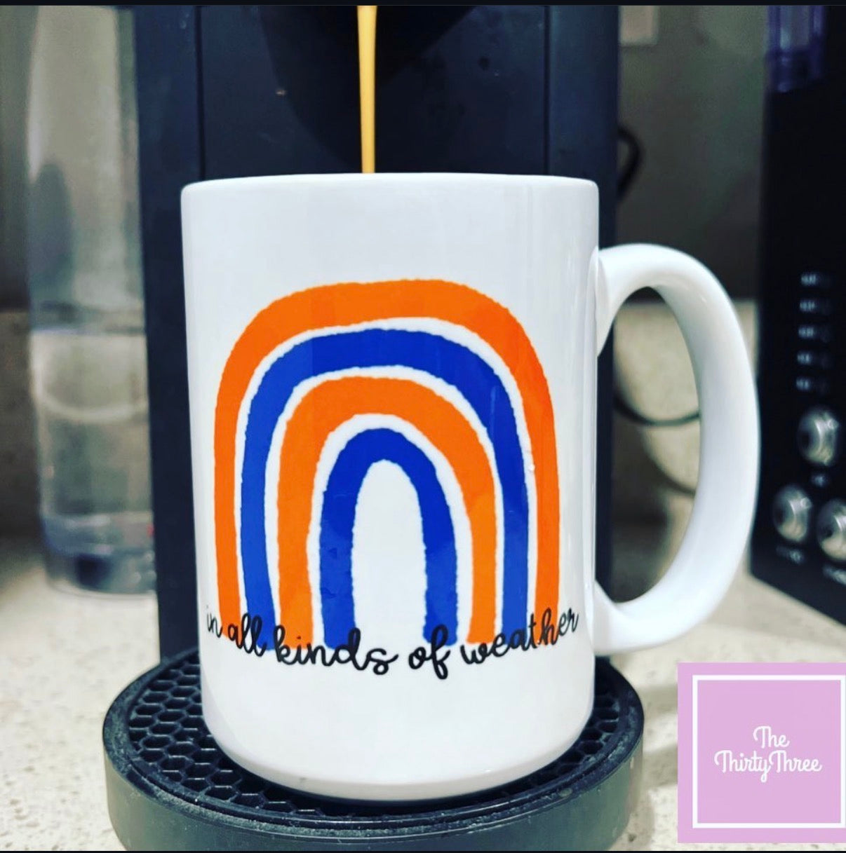 White mug with rainbow design and text on a coaster, with a blurred coffee machine in the background.